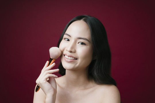 美丽的年轻亚洲女孩的画像，用化妆刷自然化妆，隔离在红色背景上。