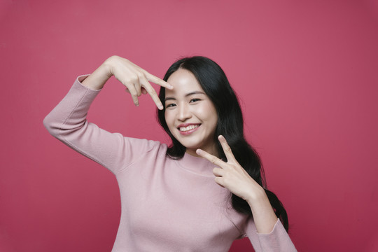 一位年轻的亚洲女性的肖像，在粉红色背景上展示了两个手指的胜利姿势。