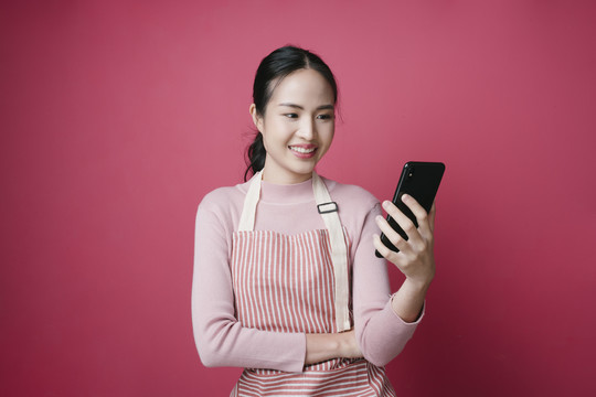 穿着围裙、使用智能手机在线购物的年轻亚洲女性肖像，粉色背景。
