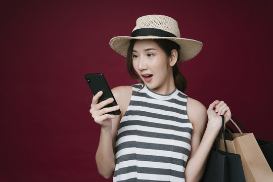 亚洲年轻女子的肖像，手持纸袋，手持智能手机，享受网上购物。