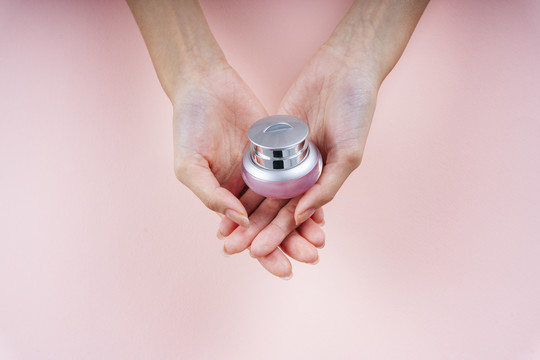 手持粉红色背景上隔离的小罐子面霜乳液的女人的手。