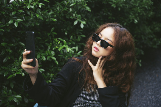 戴着墨镜和黑色夹克的亚洲女人在公园用智能手机自拍。