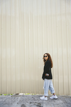 侧视图-年轻亚洲女性戴着墨镜和黑色夹克在街上行走的时尚肖像。