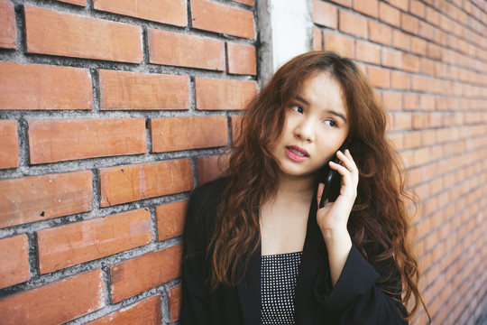 穿着黑色夹克的年轻亚洲女性在砖墙边打电话的时尚肖像。