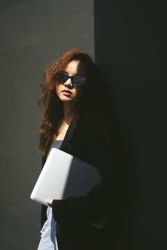 年轻亚洲女性戴着墨镜和黑色夹克手持笔记本电脑的时尚肖像。