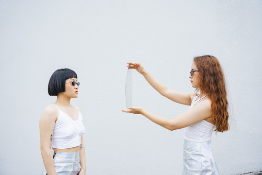 两位戴太阳镜的年轻亚洲女性的肖像。一个拿着镜子，另一个看着镜子。