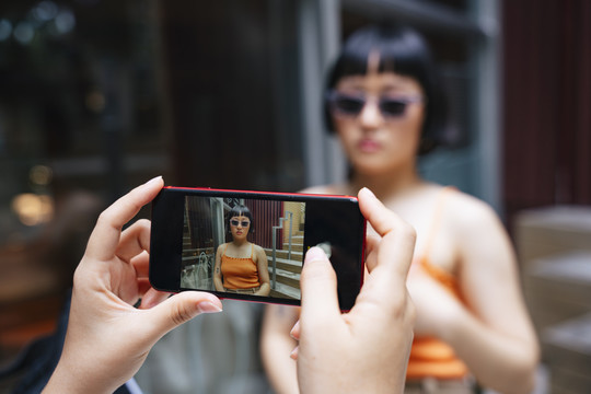 亚洲年轻女性戴着太阳镜，在咖啡馆被朋友用智能手机拍照。