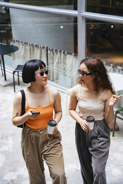 两个年轻的亚洲女孩戴着墨镜，拿着一杯咖啡在购物街上散步。