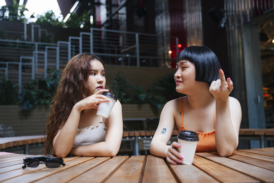 两位年轻亚洲女性在户外咖啡馆进行有趣对话的照片。