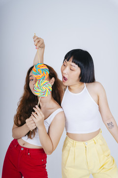 两位身穿白色背心、手持彩虹糖果棒棒糖的亚洲年轻女性的肖像，背景为白色。