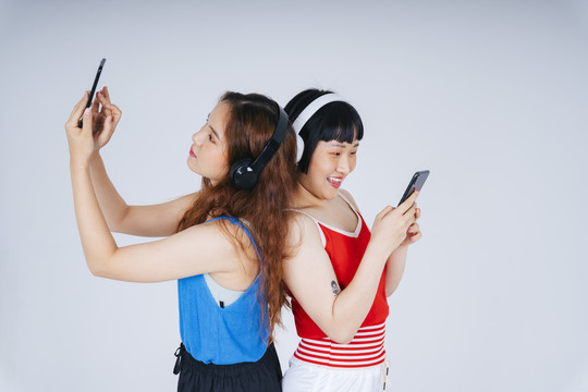 两位年轻亚洲女性在白色背景下听音乐和玩手机游戏的肖像。