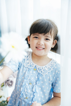 身着蓝色连衣裙的可爱小女孩在窗帘前手持白色花朵的肖像。