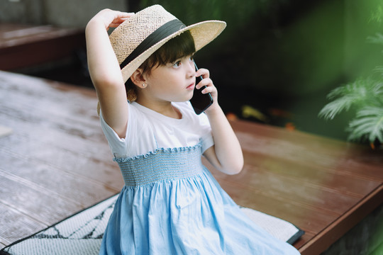 穿着白蓝色连衣裙、戴着帽子的小女孩在家里打电话。