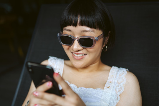 戴着墨镜的亚洲短发女性微笑着使用智能手机的特写照片。