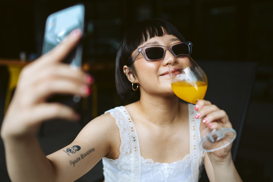戴墨镜的亚洲短发女子用橙汁自拍。