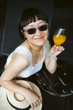戴墨镜的亚洲短发女人在沙滩上享受橙汁。