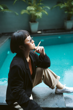 游泳池附近戴太阳镜的短发亚洲时髦女性肖像。她像香烟一样拿着钢笔。