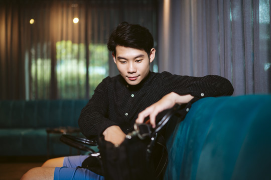 一个穿着黑衬衫的亚洲人坐在沙发上，在包里找东西。