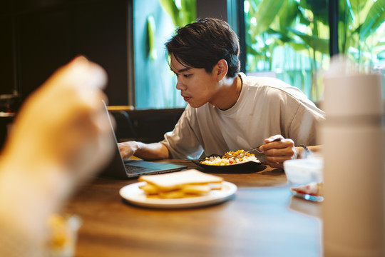 年轻的亚洲自由职业者，一边用笔记本电脑一边吃午饭。