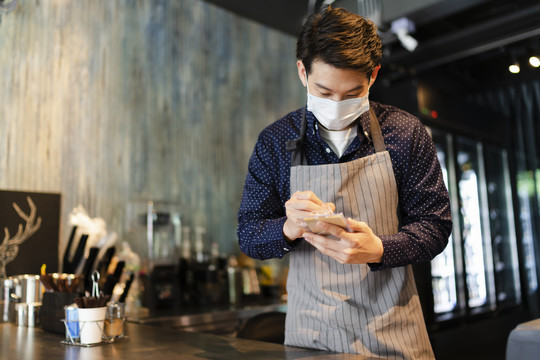 一名戴着面具的亚洲咖啡师在咖啡馆向顾客点菜。2019冠状病毒疾病概念后重新开放业务。