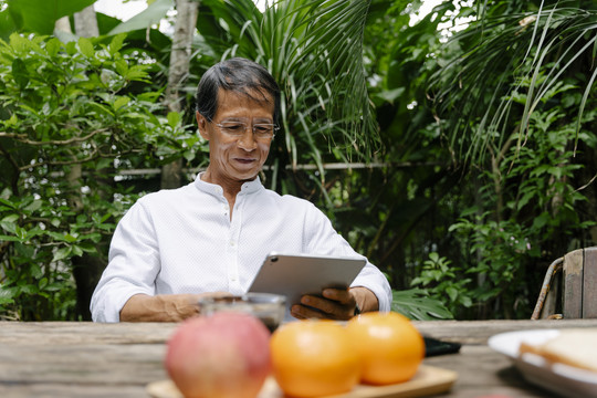 一位穿着白色休闲衬衫、戴着眼镜的亚洲老人在户外院子里使用平板电脑。