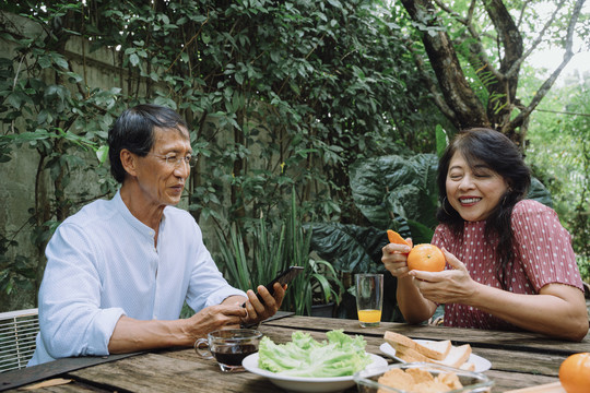 一对亚洲老夫妇退休后在后院过着美好的生活。男人用智能手机，女人剥橘皮。