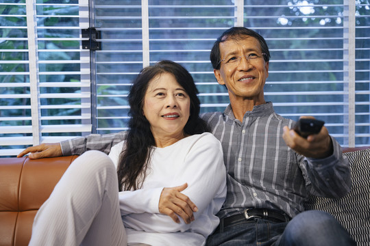 一对亚洲老年夫妇坐在沙发上，一起在家看电视。