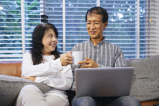 快乐的亚洲老年夫妇坐在沙发上，用数字笔记本电脑浏览网站。