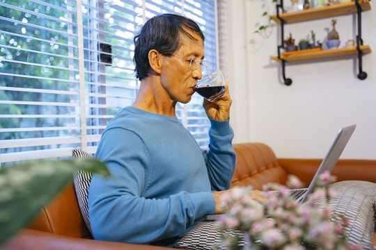 亚洲老人在家里用lapop电脑工作，端着一杯咖啡。