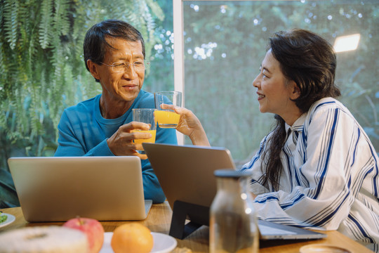亚洲的一对老年夫妇在使用笔记本电脑时喜欢喝orage果汁。