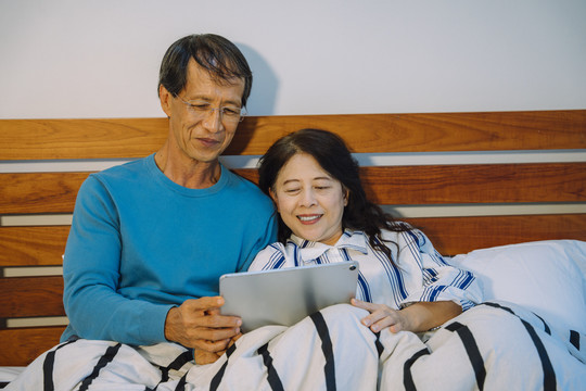 亚洲老年夫妇晚上在卧室里用数字平板电脑阅读新闻。