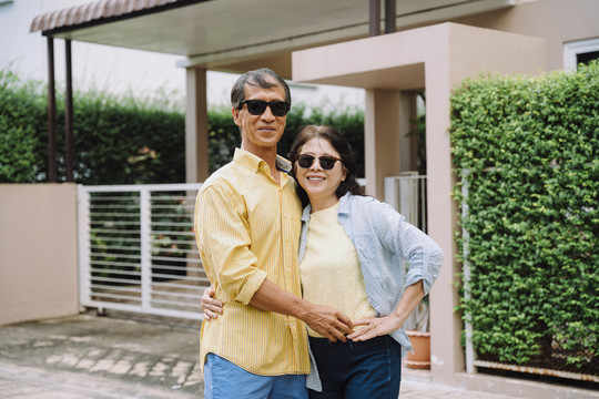 幸福的亚洲夫妇戴着墨镜站在房子前面的肖像。