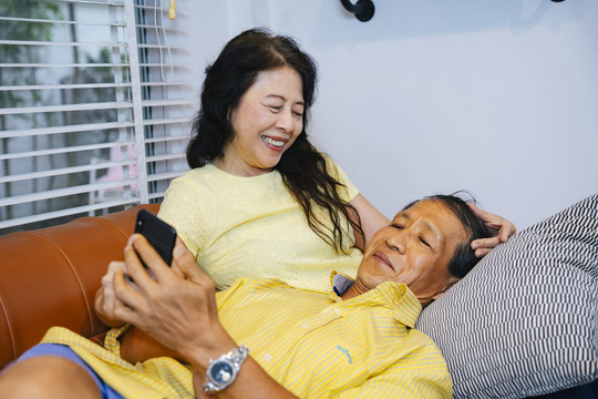 一对亚洲长辈躺在沙发上，手里拿着数字智能手机。