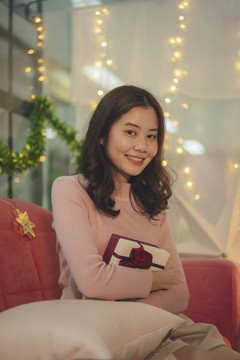 在圣诞节活动中，一位年轻的亚洲女士坐在沙发上，穿着带礼品盒的毛衣。