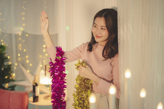 穿着粉色毛衣的亚洲年轻女子在房间里装饰圣诞道具时向某人挥手。