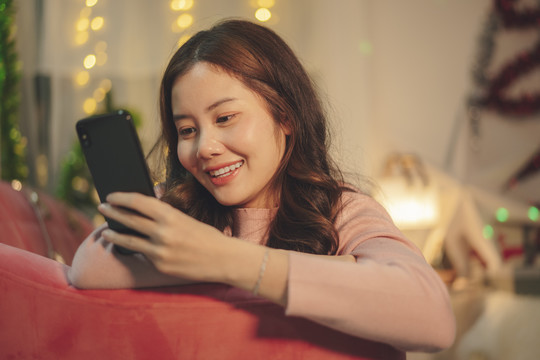 在圣诞节，年轻的亚洲美女在沙发上用智能手机梳理黑发。