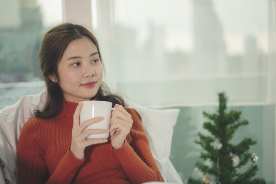 年轻的亚洲美女，黑发，穿着红色毛衣，在家里的沙发上喝着热咖啡。