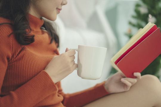 侧视图-一名妇女穿着红色毛衣看书，手里拿着热咖啡杯。