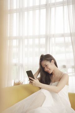 年轻的亚洲美女穿着白色衣服，在客厅的沙发上使用智能手机。