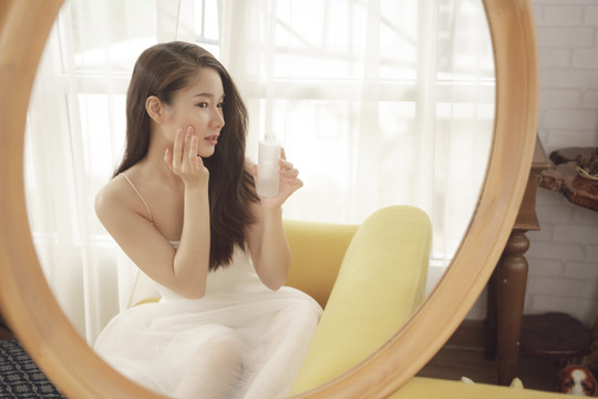 年轻的亚洲美女在镜子前使用面霜。护肤概念。
