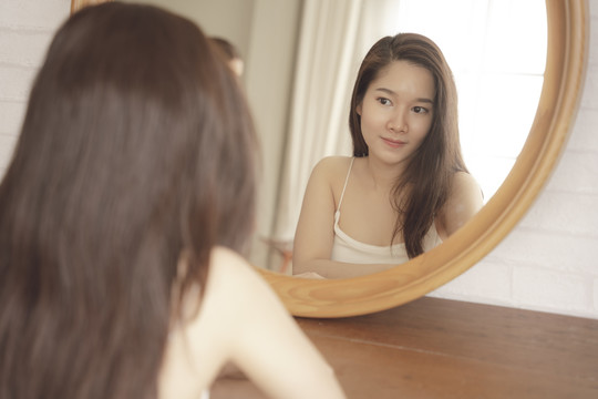 年轻的亚洲美女在镜子前的脸上涂抹护肤霜。