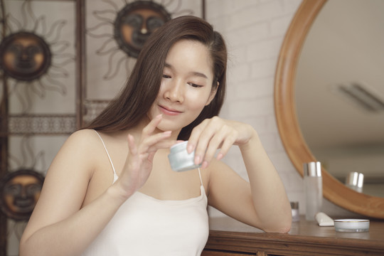 年轻的亚洲美女拿着护肤霜罐坐在化妆台上。