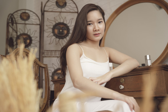 穿着白色连衣裙的年轻亚洲美女坐在化妆台旁。