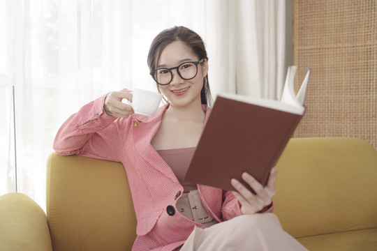 年轻的亚洲美女，穿着粉色夹克，戴着眼镜看书，喝着咖啡。
