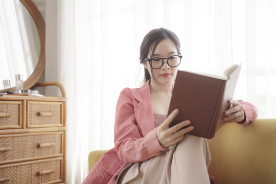 年轻的亚洲美女戴着眼镜在沙发上看书。