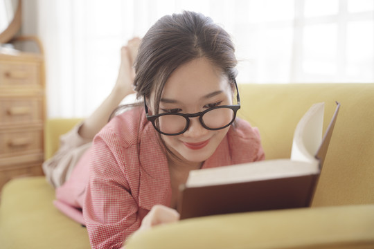 年轻的亚洲美女戴着眼镜躺在沙发上看书。