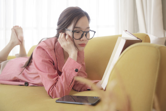 年轻的亚洲美女戴着眼镜躺在沙发上看书。