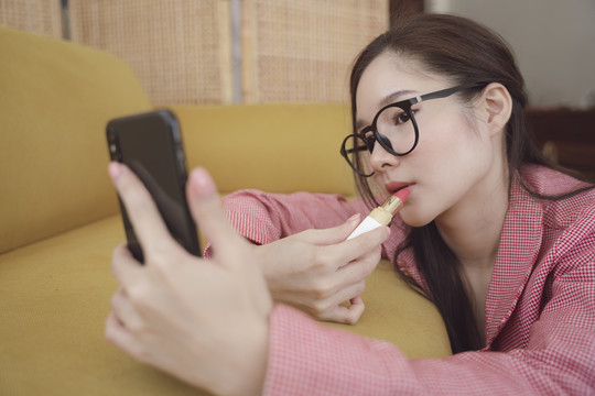 年轻的亚洲美貌女子在脸上涂口红，看着智能手机。