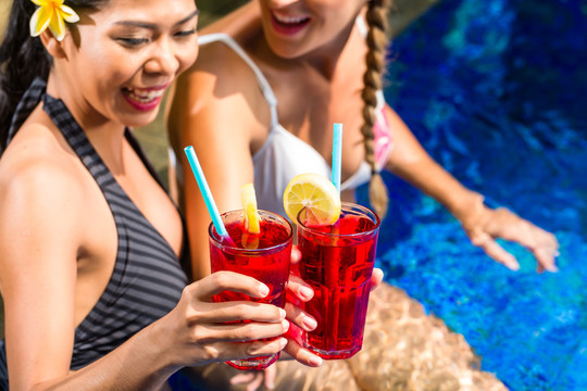 度假的两个女孩或女人，亚洲人和白种人，在酒店泳池的热带花园里晒太阳，喝饮料或鸡尾酒