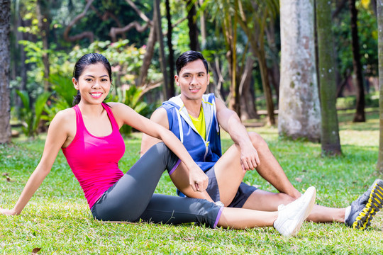 一对夫妇在热带亚洲公园做仰卧起坐，一男一女坐在草坪上做运动锻炼